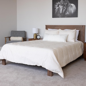 May include: A wooden bed frame with a light-colored striped comforter and matching pillows. Two wooden nightstands with gold-based lamps flank the bed. A gray armchair sits to the left, and a black and white horse print hangs on the wall.
