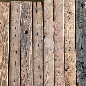 Puede incluir: Colecci&oacute;n de tablones de madera reciclada en tonos marrones y grises. Los tablones presentan vetas, nudos y agujeros de clavos visibles, mostrando su aspecto r&uacute;stico y envejecido. La madera est&aacute; dispuesta verticalmente, creando una superficie texturizada.