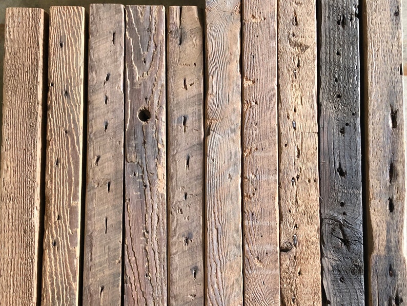 Puede incluir: Colecci&oacute;n de tablones de madera reciclada en tonos marrones y grises. Los tablones presentan vetas, nudos y agujeros de clavos visibles, mostrando su aspecto r&uacute;stico y envejecido. La madera est&aacute; dispuesta verticalmente, creando una superficie texturizada.
