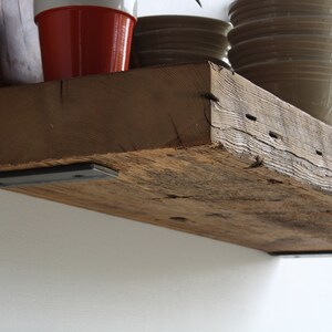 Puede incluir: Estanter&iacute;a r&uacute;stica de madera reciclada con vetas y agujeros de clavos visibles. Est&aacute; soportada por dos soportes met&aacute;licos oscuros y montada en una pared blanca. Varios objetos, como cuencos y una maceta roja, se exhiben en la estanter&iacute;a. La profundidad de la estanter&iacute;a es de unos 20 cm.