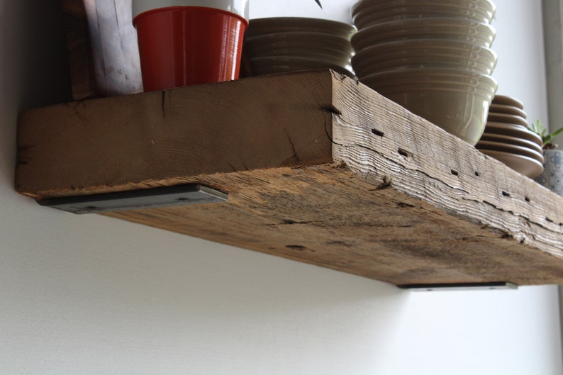 Puede incluir: Estanter&iacute;a r&uacute;stica de madera reciclada con vetas y agujeros de clavos visibles. Est&aacute; soportada por dos soportes met&aacute;licos oscuros y montada en una pared blanca. Varios objetos, como cuencos y una maceta roja, se exhiben en la estanter&iacute;a. La profundidad de la estanter&iacute;a es de unos 20 cm.