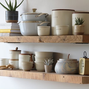 May include: Two rustic wooden shelves display kitchenware and decor. Items include light gray pots, cream-colored canisters labeled "FLOUR" and "PASTA", and small ceramic bowls. A small succulent and a potted plant add greenery.