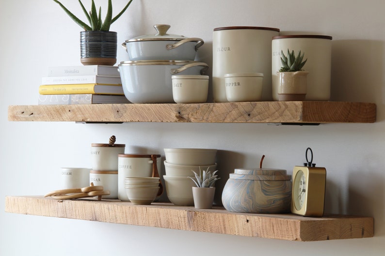 May include: Two rustic wooden shelves display kitchenware and decor. Items include light gray pots, cream-colored canisters labeled "FLOUR" and "PASTA", and small ceramic bowls. A small succulent and a potted plant add greenery.