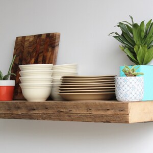 Puede incluir: Una estanter&iacute;a r&uacute;stica de madera exhibe una colecci&oacute;n de objetos. Hay pilas de cuencos blancos y platos color canela, una tabla de cortar de madera y plantas en macetas. Las plantas incluyen aloe vera y una planta de hojas verdes en una maceta turquesa.