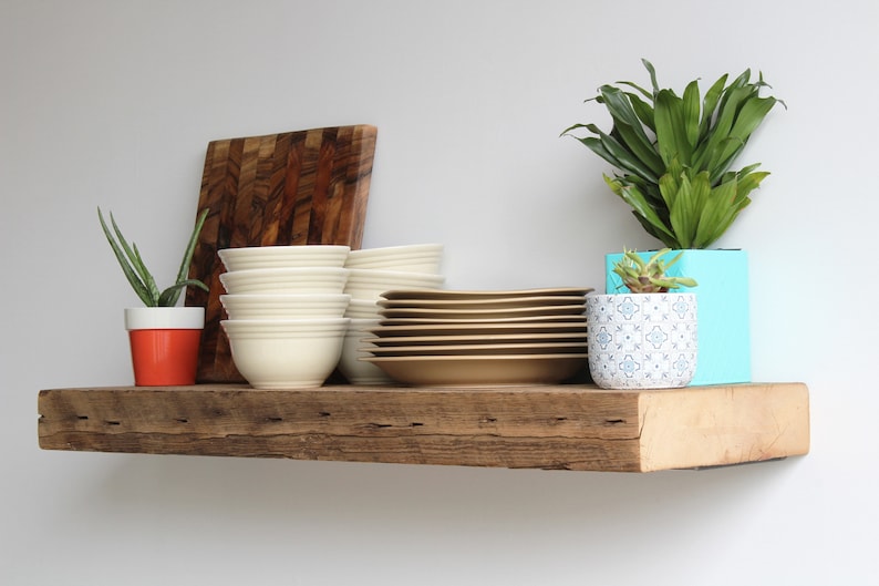 Puede incluir: Una estanter&iacute;a r&uacute;stica de madera exhibe una colecci&oacute;n de objetos. Hay pilas de cuencos blancos y platos color canela, una tabla de cortar de madera y plantas en macetas. Las plantas incluyen aloe vera y una planta de hojas verdes en una maceta turquesa.