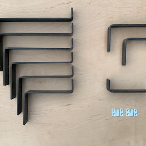 May include: A collection of dark gray metal shelf brackets in various sizes, arranged on a light wood surface. The brackets are L-shaped, with pre-drilled holes for mounting. Also included are several small, white metal mounting plates.