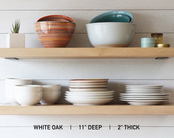 May include: Two wooden shelves displaying kitchenware. The top shelf has bowls in orange, teal, and white. The bottom shelf shows white bowls and plates. Text at the bottom reads: WHITE OAK | 11" DEEP | 2" THICK.