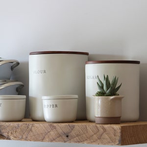 May include: A kitchen shelf displays a collection of off-white canisters labeled "FLOUR", "SUGAR", "SALT", and "PEPPER", each with a dark wooden lid. A small potted succulent sits next to the sugar canister. Light blue cookware is stacked on the left.