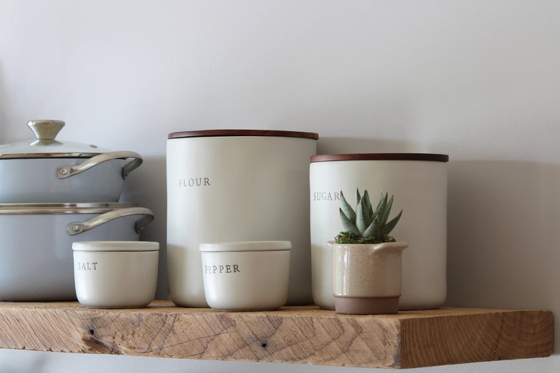 May include: A kitchen shelf displays a collection of off-white canisters labeled "FLOUR", "SUGAR", "SALT", and "PEPPER", each with a dark wooden lid. A small potted succulent sits next to the sugar canister. Light blue cookware is stacked on the left.