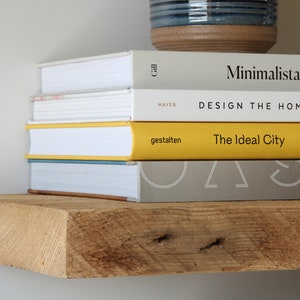 May include: A stack of books on a rustic wooden shelf. The books have white, yellow, and gray covers with titles like "Minimalista" and "The Ideal City." A blue and brown ceramic pot sits above the books.