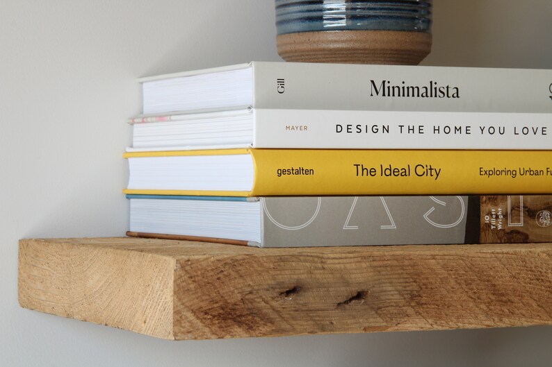 May include: A stack of books on a rustic wooden shelf. The books have white, yellow, and gray covers with titles like "Minimalista" and "The Ideal City." A blue and brown ceramic pot sits above the books.
