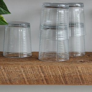 May include: Clear, ribbed drinking glasses are stacked and arranged on a rustic, wooden shelf. The glasses are inverted, and the shelf has a natural, weathered appearance. A green plant is visible in the background.