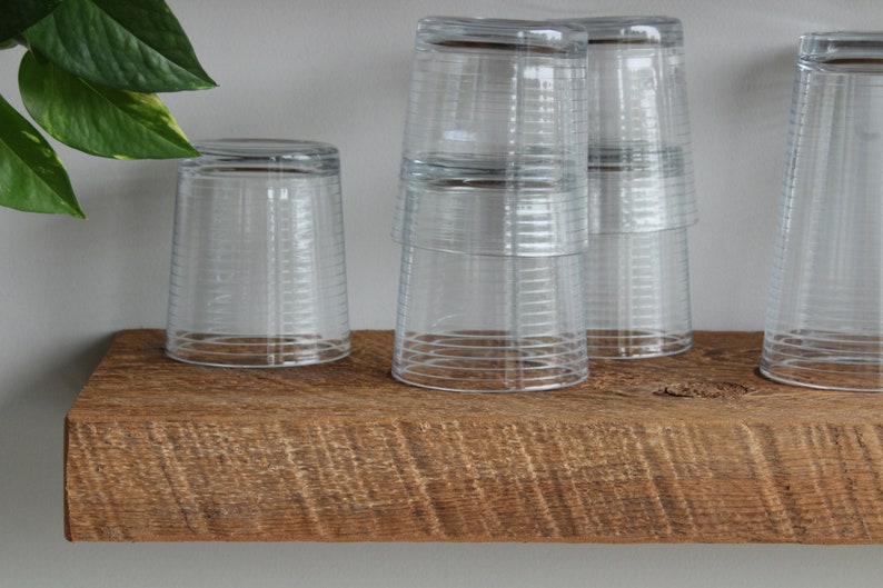 May include: Clear, ribbed drinking glasses are stacked and arranged on a rustic, wooden shelf. The glasses are inverted, and the shelf has a natural, weathered appearance. A green plant is visible in the background.