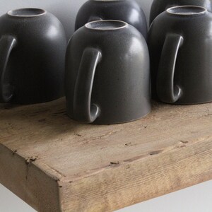 May include: A collection of dark gray ceramic mugs with handles, resting upside down on a rustic, weathered wooden shelf. The mugs have a speckled texture and a simple, modern design, perfect for coffee or tea.