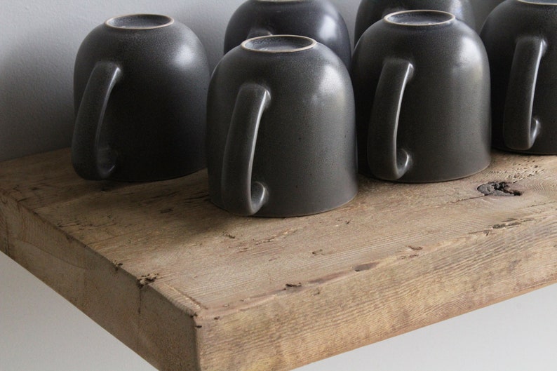 May include: A collection of dark gray ceramic mugs with handles, resting upside down on a rustic, weathered wooden shelf. The mugs have a speckled texture and a simple, modern design, perfect for coffee or tea.