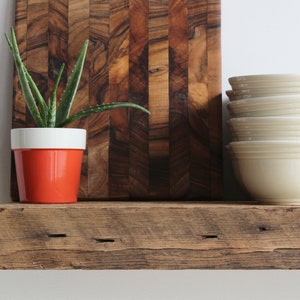 Puede incluir: Una estanter&iacute;a r&uacute;stica de madera exhibe una peque&ntilde;a planta de aloe vera en una maceta naranja y blanca, una pila de cuencos color crema y una tabla de cortar de madera. La tabla de cortar tiene un patr&oacute;n de rayas verticales marr&oacute;n oscuro. La estanter&iacute;a es de madera envejecida.