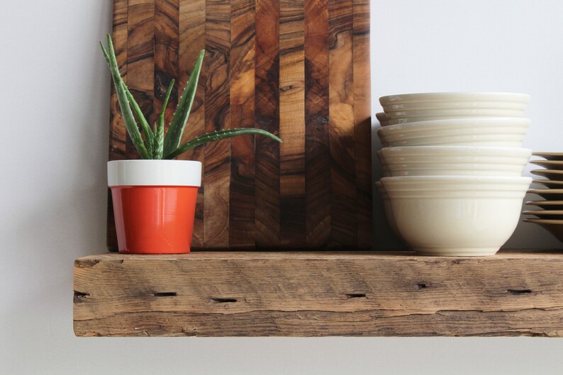 Puede incluir: Una estanter&iacute;a r&uacute;stica de madera exhibe una peque&ntilde;a planta de aloe vera en una maceta naranja y blanca, una pila de cuencos color crema y una tabla de cortar de madera. La tabla de cortar tiene un patr&oacute;n de rayas verticales marr&oacute;n oscuro. La estanter&iacute;a es de madera envejecida.