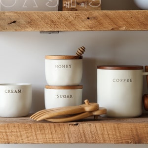 May include: A collection of white ceramic kitchen canisters and serving dishes with wooden lids and accents. The canisters are labeled "HONEY", "SUGAR", and "COFFEE". A wooden honey dipper and serving utensils are also visible.