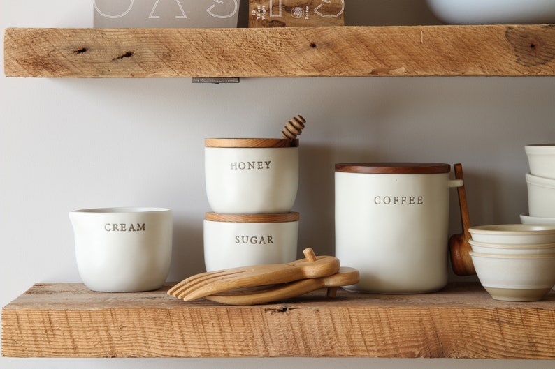 May include: A collection of white ceramic kitchen canisters and serving dishes with wooden lids and accents. The canisters are labeled "HONEY", "SUGAR", and "COFFEE". A wooden honey dipper and serving utensils are also visible.