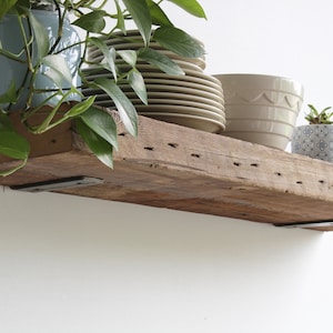 Puede incluir: Una estanter&iacute;a r&uacute;stica de madera con soportes met&aacute;licos, que sostiene varios objetos. Hay pilas de platos color beige, un cuenco beige y una maceta peque&ntilde;a con un estampado y una planta. Una cesta de alambre contiene manzanas rojas.