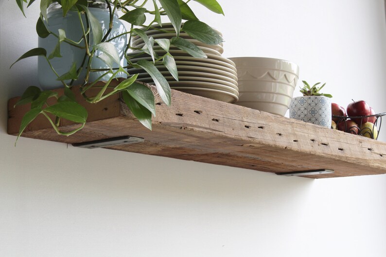 Puede incluir: Una estanter&iacute;a r&uacute;stica de madera con soportes met&aacute;licos, que sostiene varios objetos. Hay pilas de platos color beige, un cuenco beige y una maceta peque&ntilde;a con un estampado y una planta. Una cesta de alambre contiene manzanas rojas.
