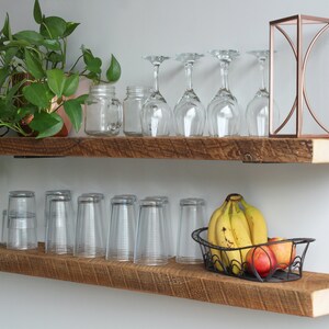 May include: Two rustic wooden shelves mounted on a white wall. The top shelf holds a potted plant, glass jars, wine glasses, and a copper geometric decoration. The bottom shelf displays drinking glasses and a wire basket of fruit.