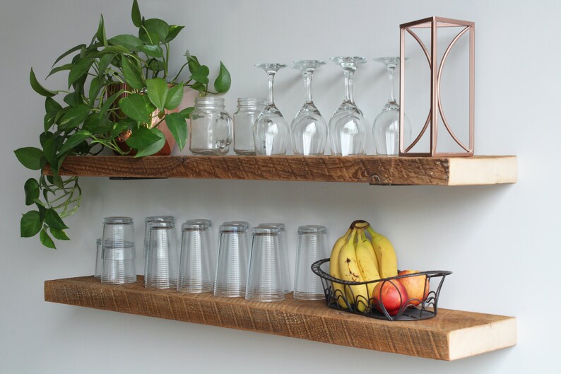 May include: Two rustic wooden shelves mounted on a white wall. The top shelf holds a potted plant, glass jars, wine glasses, and a copper geometric decoration. The bottom shelf displays drinking glasses and a wire basket of fruit.