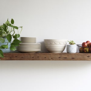 Puede incluir: Una estanter&iacute;a r&uacute;stica de madera exhibe varios objetos contra una pared blanca. Una planta en maceta, una pila de platos, un cuenco grande de color crema, una peque&ntilde;a planta suculenta en maceta y una cesta de alambre con manzanas.
