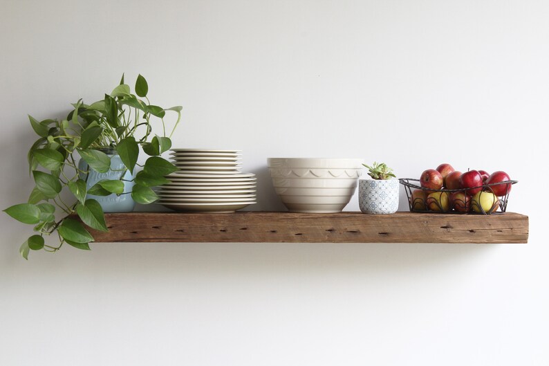 Puede incluir: Una estanter&iacute;a r&uacute;stica de madera exhibe varios objetos contra una pared blanca. Una planta en maceta, una pila de platos, un cuenco grande de color crema, una peque&ntilde;a planta suculenta en maceta y una cesta de alambre con manzanas.