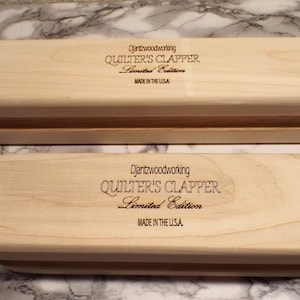 May include: Two wooden Quilter's Clappers, rectangular with rounded edges, are displayed. The text "Djantzwoodworking QUILTER'S CLAPPER Limited Edition MADE IN THE USA" is engraved on the top. The clappers are made of light-colored wood.