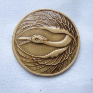 May include: A circular, beige decorative item featuring a carved bird motif. The bird is shown in profile, with an open beak and outstretched wings. The item has a raised edge and is set against a white backdrop.