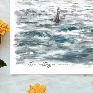 May include: Watercolor print of a lake scene with a person swimming. The artwork features shades of blue and gray, with the text "the cottage" and "Lake Muskoka" in the lower portion. Yellow flowers are in the foreground.