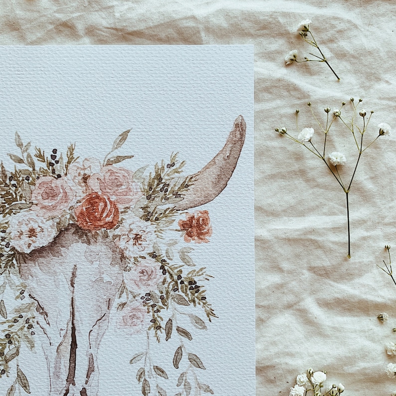 May include: A watercolor painting of a white animal skull with a floral crown of pink and red flowers. The background is a light beige.