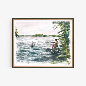 May include: Watercolor painting of a lake scene with two people. One person is sitting on a rock, and another is diving into the water. The painting is framed in a dark green frame. The text "the cottage" is written at the bottom.