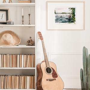 May include: Acoustic guitar with a natural wood finish, leaning against a white wall. A framed watercolor painting of a lake scene hangs nearby. A white bookshelf is filled with books and decorative items.
