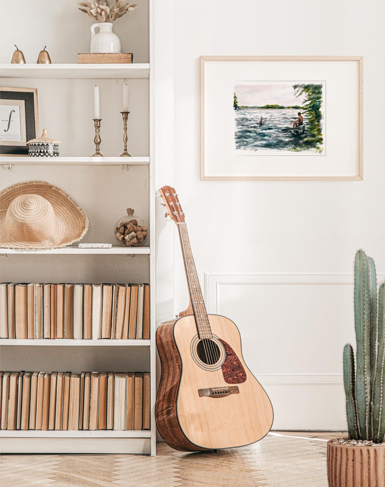 May include: Acoustic guitar with a natural wood finish, leaning against a white wall. A framed watercolor painting of a lake scene hangs nearby. A white bookshelf is filled with books and decorative items.
