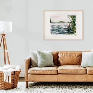 May include: A framed watercolor painting depicts a serene lake scene. A person sits on a rock, while another swims in the water. The artwork features shades of blue, green, and brown, with a light-colored mat and a natural wood frame.
