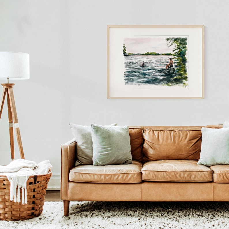May include: A framed watercolor painting depicts a serene lake scene. A person sits on a rock, while another swims in the water. The artwork features shades of blue, green, and brown, with a light-colored mat and a natural wood frame.