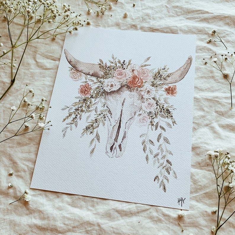 May include: A watercolor painting of a white animal skull with a floral arrangement of pink, white, and red flowers. The skull has horns and is surrounded by green leaves.