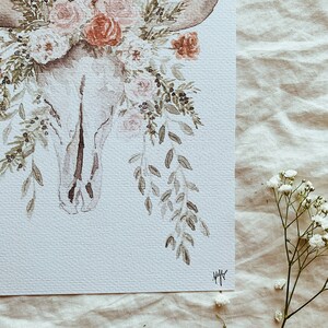 May include: A watercolor painting of a white animal skull with delicate floral accents. The skull is adorned with pink and red roses, green leaves, and small berries. The painting has a soft, romantic feel.