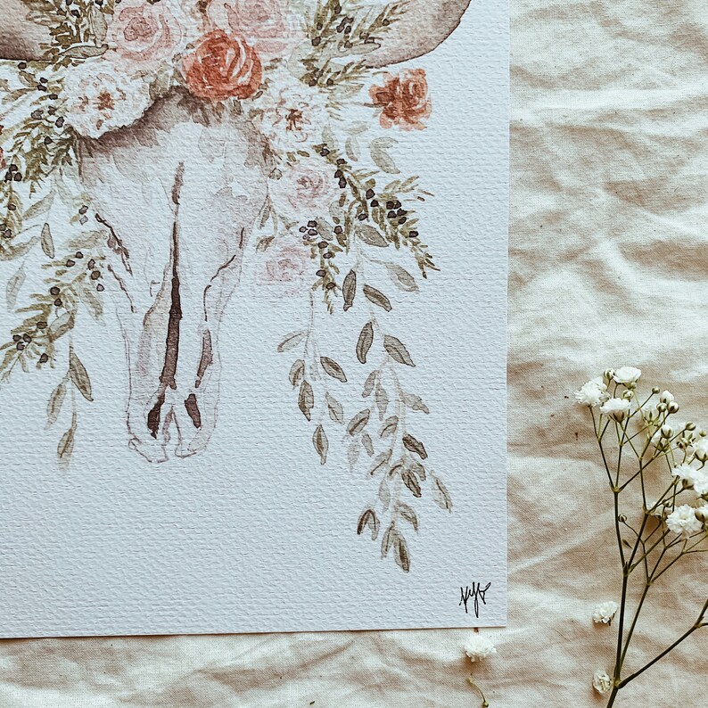 May include: A watercolor painting of a white animal skull with delicate floral accents. The skull is adorned with pink and red roses, green leaves, and small berries. The painting has a soft, romantic feel.