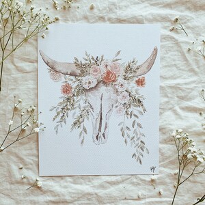 May include: A watercolor painting of a white animal skull with a floral crown of pink and orange flowers. The skull is surrounded by green leaves and vines.