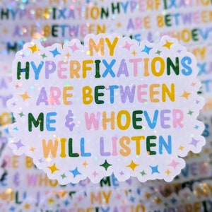 Op de afbeelding: Een witte sticker in de vorm van een wolk met een glinsterende afwerking. De sticker heeft de tekst "Mijn hyperfixaties liggen tussen mij en wie dan ook die wil luisteren" in kleurrijke letters.