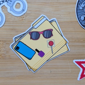 May include: A yellow file folder sticker featuring a blue smartphone, pink-framed sunglasses, a purple nail polish bottle, and a red lollipop. The sticker has a white border and is designed with a flat, graphic illustration style.