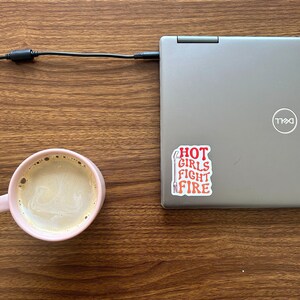 May include: A pink ceramic mug filled with coffee sits on a wooden table next to a silver laptop with a sticker that reads "HOT GIRLS FIGHT FIRE".