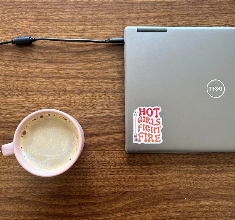 May include: A pink ceramic mug filled with coffee sits on a wooden table next to a silver laptop with a sticker that reads "HOT GIRLS FIGHT FIRE".
