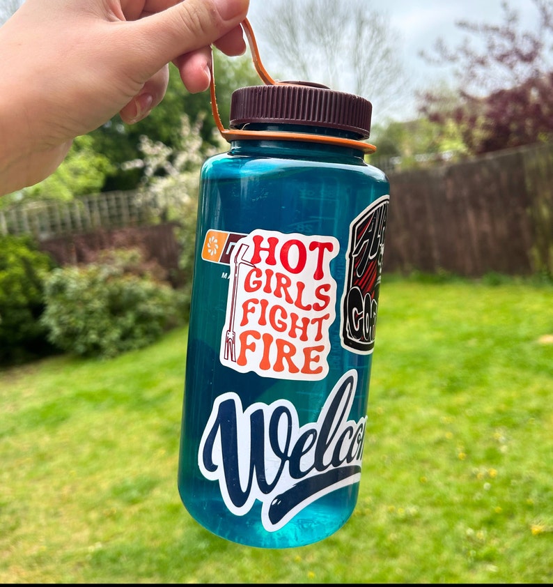 May include: A blue reusable water bottle with a brown lid and a carrying handle. The bottle has several stickers on it, including one that says "Hot Girls Fight Fire".