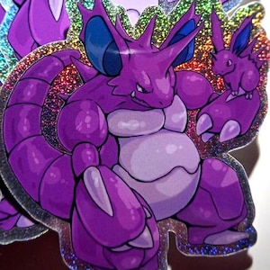 May include: A stack of holographic stickers featuring a purple Pokemon character with blue eyes and a smaller purple Pokemon character with blue eyes. The stickers have a glittery finish.