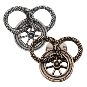 May include: Two metal pins featuring a wheel design intertwined with a snake. One pin is silver-toned, the other is bronze-toned. The snake is coiled around the wheel, creating a unique and detailed design. These pins are likely decorative accessories.