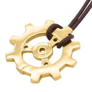 Könnte beinhalten: Ein goldfarbener Zahnrad-Anhänger mit einem braunen Lederband. Das Zahnrad hat ein detailliertes Design mit einer zentralen Nabe und Speichen.